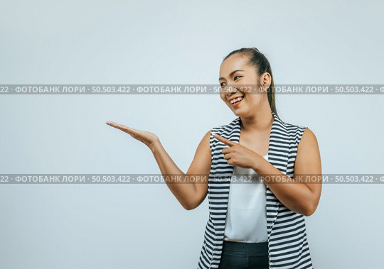 Portrait of Asian woman over isolated background in studio with blank space. Smiling aside while presenting with hand and pointing with finger. Стоковое фото, агентство Ingram Publishing / Фотобанк Лори