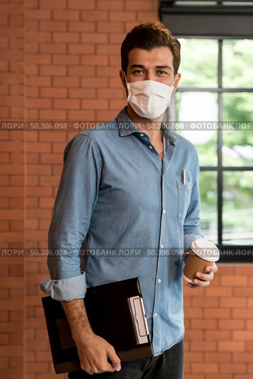 Portrait of middle east caucasian businessman with takeout coffee cup and clipboard in meeting room after lunch break. He wear face mask as new normal office preventing coronavirus COVID-19 spreading. Стоковое фото, агентство Ingram Publishing / Фотобанк Лори