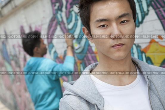 Portrait of serious young man with a cool attitude looking at the camera, young man spray painting in the background. Стоковое фото, агентство Ingram Publishing / Фотобанк Лори