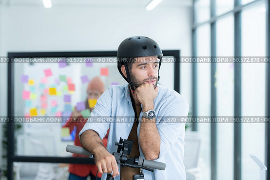 Portrait of smiling businessman riding electric scooter in office with colleague in background. Стоковое фото, агентство Ingram Publishing / Фотобанк Лори