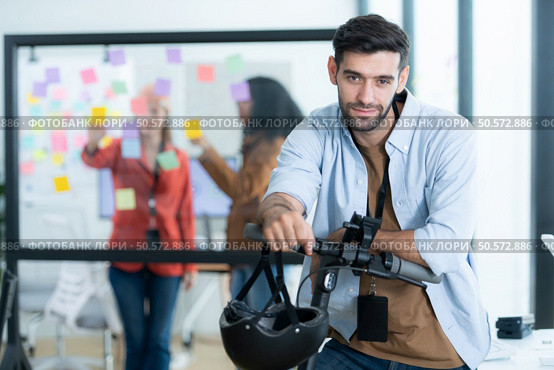 Portrait of smiling businessman riding electric scooter in office with colleague in background. Стоковое фото, агентство Ingram Publishing / Фотобанк Лори