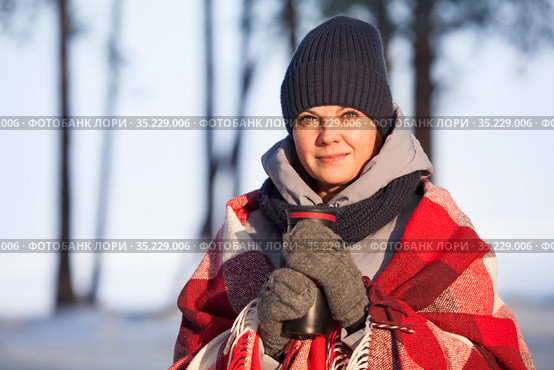 Portrait of young beautiful woman in winter clothes, red scarf and mittens holding thermo cup with hot beverage standing in sunny cold forest. Стоковое фото, фотограф Кекяляйнен Андрей / Фотобанк Лори