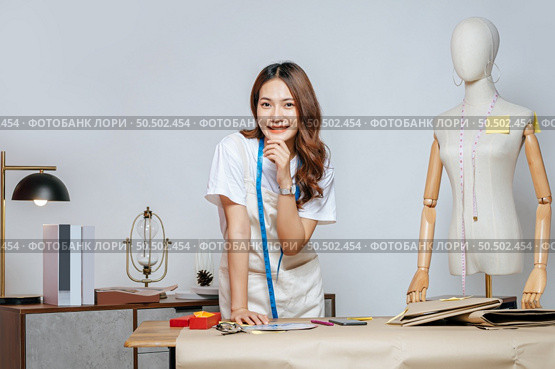 Portrait of young pretty female fashion designer stylish standing and hand on shin with smile feeling proud her business during working at fashion studio, full of tailoring tools and equipment. Стоковое фото, агентство Ingram Publishing / Фотобанк Лори