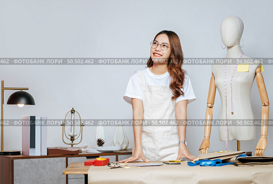 Portrait of young pretty female fashion designer stylish wearing eyeglasses standing and smile with proud during working at fashion studio, full of tailoring tools and equipment. Стоковое фото, агентство Ingram Publishing / Фотобанк Лори