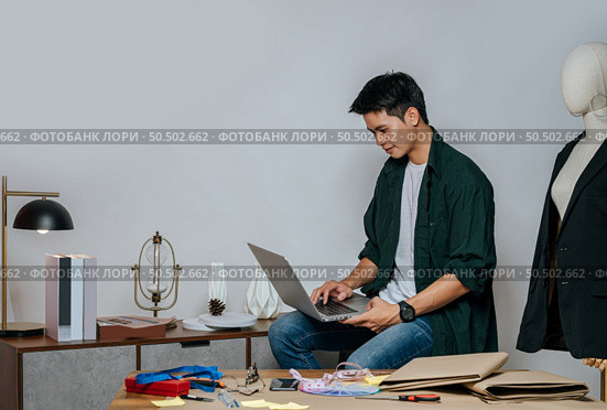 Portrait Young handsome male designer use laptop computer for searching good idea to design fashion clothing, mannequin and equipment with full of tailoring tools on desk in studio. Стоковое фото, агентство Ingram Publishing / Фотобанк Лори
