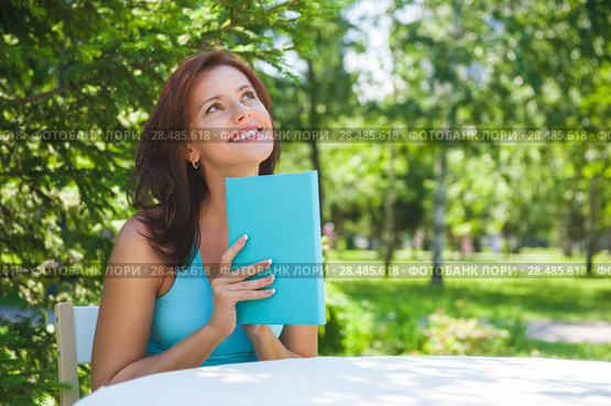 Positive adult beautiful woman sitting near table and reading book outdoors at backyard in summer. Стоковое фото, фотограф Kirill Kedrinskiy / Ingram Publishing / Фотобанк Лори