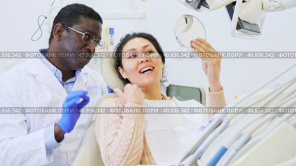 Positive african american dentist talking to asian woman during dental checkup, discussing teeth x-ray and treatment procedures. Стоковое видео, видеограф Яков Филимонов / Фотобанк Лори