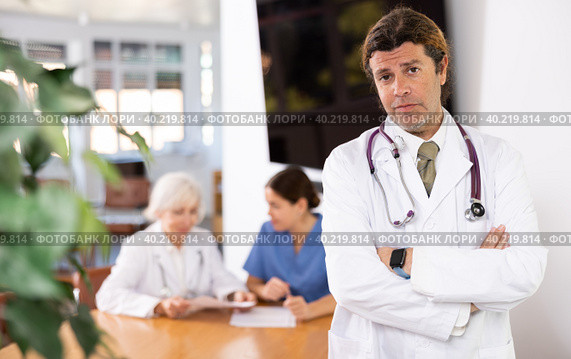 positive medical male colleague standing in meeting room and employees are talking behind him at table. Стоковое фото, фотограф Яков Филимонов / Фотобанк Лори