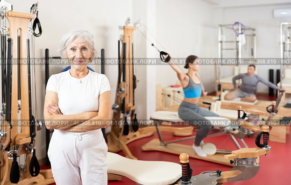 Positive senior woman coach stands in large gym against background of Pilates simulators. Стоковое фото, фотограф Яков Филимонов / Фотобанк Лори