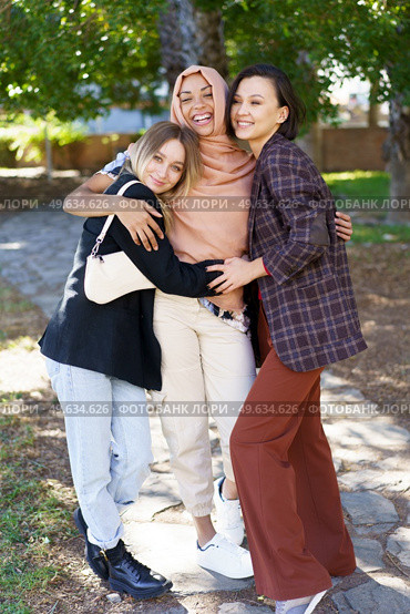 Positive young multiethnic female best friends in casual clothes smiling and looking at camera while cuddling in park on sunny day. Delighted young diverse ladies embracing and smiling in park. Стоковое фото, агентство Ingram Publishing / Фотобанк Лори