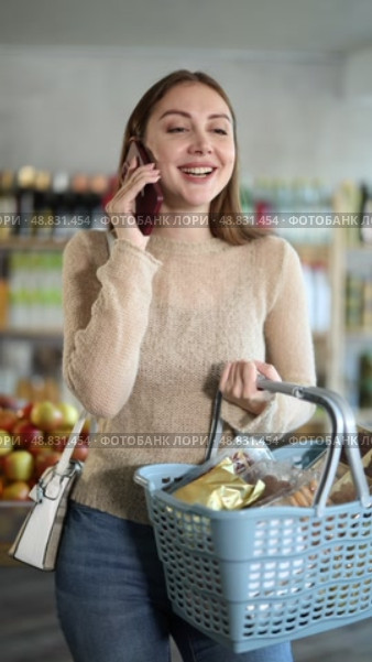 Positive young woman looking for something, holding cart with food products and talking on mobile phone at store. Стоковое видео, видеограф Яков Филимонов / Фотобанк Лори