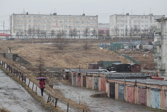 Повседневный городской пейзаж. Вид на гаражи и панельные дома. По пешеходной дорожке идёт женщина с зонтом. Пасмурная погода. Атмосферные осадки в виде мокрого снега с дождём. Город Магадан, Магаданская область, Россия. (2024 год). Редакционное фото, фотограф Андрей Степанов / Фотобанк Лори