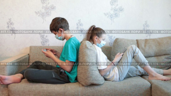 Pre-teen sister and brother gaming online with cellphones while sitting at home, children wearing safety masks, sitting back to back on sofa. Стоковое видео, видеограф Кекяляйнен Андрей / Фотобанк Лори