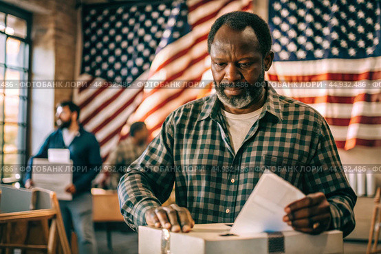 President elections. A black man votes for the new president of the United States of America and puts a form in the ballot box against the background of the American flag. Стоковое AI-изображение, AI-художник Photojuli / Фотобанк Лори