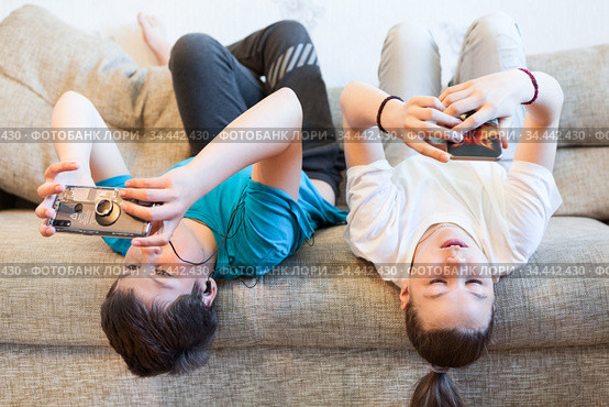 Preteen boy and girl using smartphones for on-line conversation, kids lying back on sofa, idleness. Стоковое фото, фотограф Кекяляйнен Андрей / Фотобанк Лори