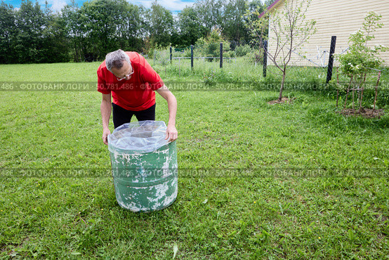 Pulling plastic liner over rim of weathered water butt standing on grassy garden lawn with open space and clear sky in background. Стоковое фото, фотограф Владимир Григорьев / Фотобанк Лори