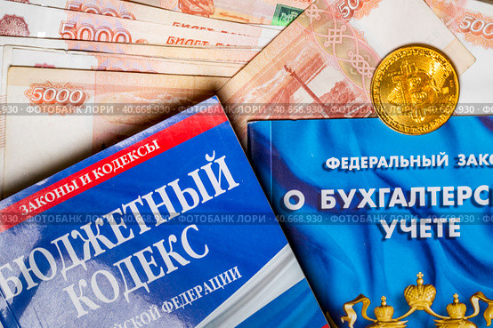 Пятитысячные купюры, биткоин и брошюры с федеральным законом Российской Федерации "О бухгалтерском учете" и Бюджетный кодекс лежат на столе у бухгалтера (2023 год). Редакционное фото, фотограф Николай Винокуров / Фотобанк Лори