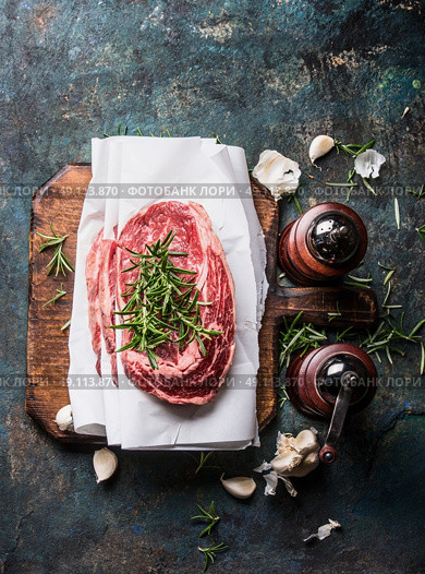 Raw marbled meat steaks with wrapping paper on rustic cutting board with fresh flavoring and spice mills on dark rustic background, top view. Стоковое фото, агентство Ingram Publishing / Фотобанк Лори