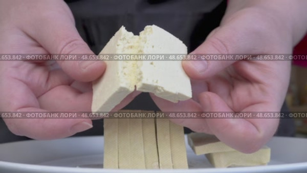 Разламывание куска тофу руками. Крупный план: человек показывает текстуру свежего тофу, разламывая кусочек руками. Видны слоистая структура и плотность продукта. Внизу на тарелке — аккуратно нарезанные ломтики. Стоковое видео, видеограф Анна Чижова / Фотобанк Лори