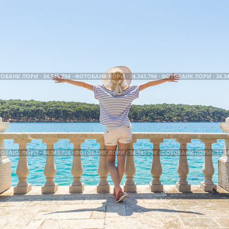 Rear view of happy woman on vacation, wearing straw summer hat ,standing on luxury elegant old stone balcony of coastal villa, relaxing, arms rised to the sun, looking at blue Adriatic sea. Стоковое фото, фотограф Matej Kastelic / Фотобанк Лори