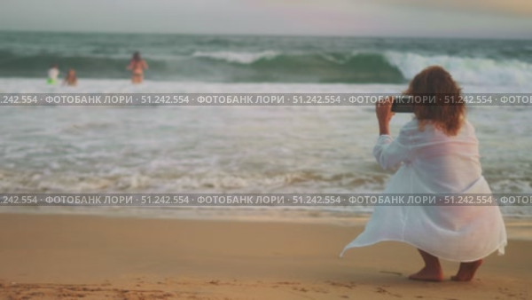 Rear view of woman filming waves and people playing in ocean at sunset. Relaxed mood, soft light, evening sea breeze. Mindful moment, focused on memories and serenity. Стоковое видео, видеограф Енваев Андрей / Фотобанк Лори