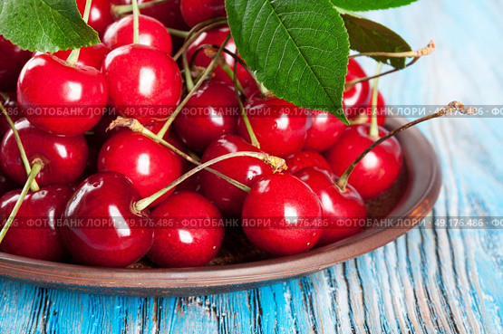 Red sweet cherry and green leaves in a clay dish on a blue wooden background. Стоковое фото, агентство Ingram Publishing / Фотобанк Лори