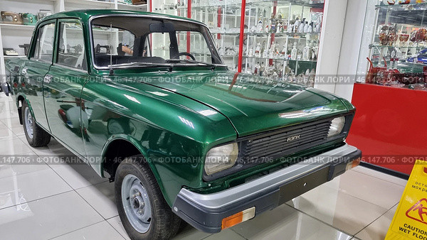 Retro car "AZLK Moskvich-2140", painted in a rich green color. This Soviet-made car was produced at the Moscow Automobile Plant (AZLK) in the 1970-1980s. The car is in the USSR Museum, surrounded by display cases with porcelain figurines, crystal and other items of Soviet life.Classic Soviet car with a characteristic design.Green body paint, shiny chrome bumper.The signature "AZLK" emblem and the "Moskvich-2140" model on the front.Museum setting, glass display cases with items from the Soviet era.Caution, wet floor! - warning sign on the floor.Retro-style museum interior with red design elements. (2023 год). Редакционное фото, фотограф Анна Кривицкая / Фотобанк Лори