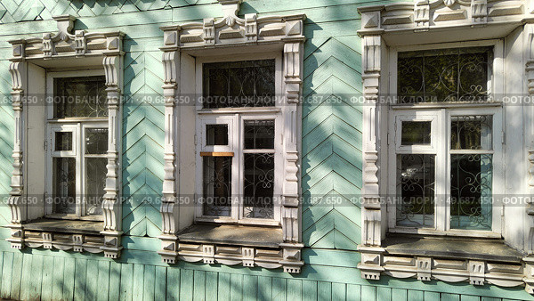 Резные наличники на окнах деревянного здания. Традиционный декор русских домов. Три окна с белыми резными наличниками на стене мятного цвета старинной усадьбы. Фасад с декоративной отделкой. Carved architraves on windows of wooden building. Traditional decor of Russian houses. Three windows with white carved platbands on mint-colored wall of old manor house. Facade with decorative trim. Стоковое фото, фотограф Ирина / Фотобанк Лори