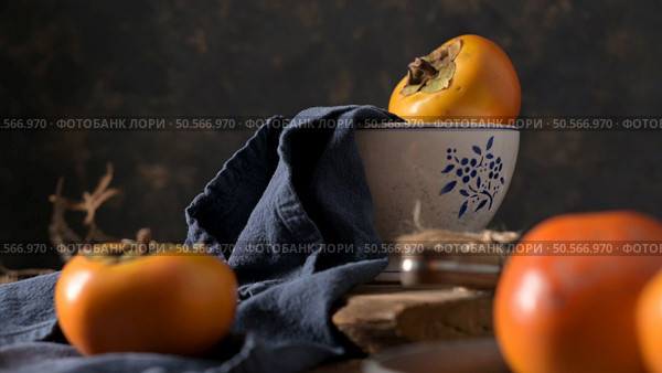 Ripe persimmons on a navy cloth with rustic wooden elements create a cozy autumn scene. Soft lighting highlights the fruit’s vibrant color and texture. Стоковое фото, агентство Ingram Publishing / Фотобанк Лори