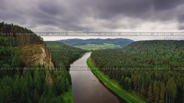 River flows through green forest near rocky cliff under cloudy sky. Стоковое AI-видео, аниматор Юрий Поздников / Фотобанк Лори