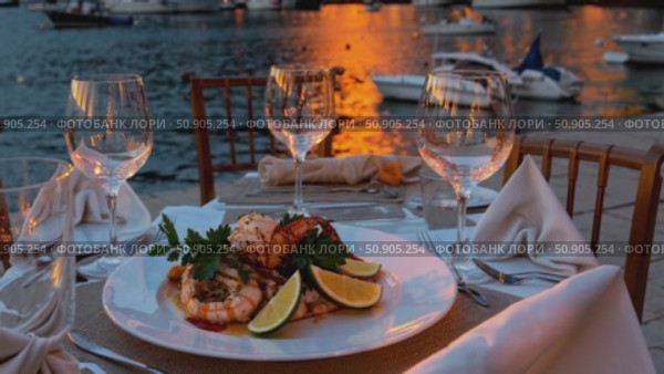 Romantic seaside dining featuring elegant seafood platter, crystal wine glasses, and candlelit table overlooking tranquil ocean waves at sunset. Стоковое AI-видео, аниматор Данил Руденко / Фотобанк Лори