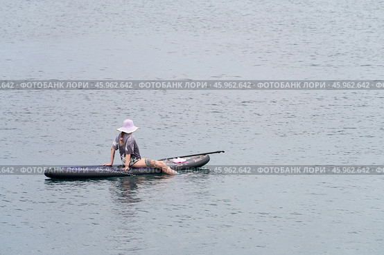 Russia, Bolshoi Utrish, September 16, 2024: a woman sits on a sapboard in a sea bay. Her feet are lowered into the water. Paddle and beach shoes on the board. Стоковое фото, фотограф Роман Дзюбало / Фотобанк Лори