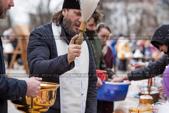 Russia, the City of Vidnoye, April 15, 2023. Easter service. The priest sprinkles people and Easter treats with holy water at the entrance to the temple. People go to sanctify eggs and cakes. Редакционное фото, фотограф Photojuli / Фотобанк Лори