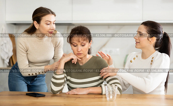 Sad woman sits silently in kitchen, adult girls sympathize and comfort her. Стоковое фото, фотограф Яков Филимонов / Фотобанк Лори