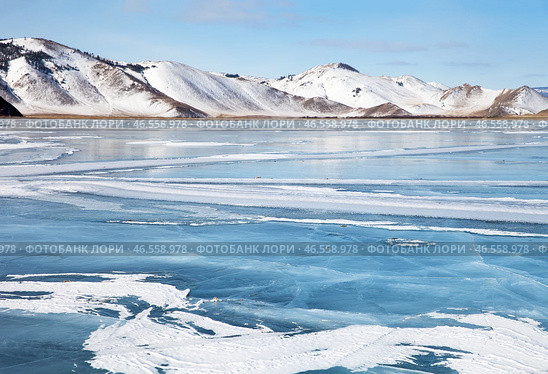 Scenic winter landscape of frozen Baikal lake on cold sunny day in February. Snow-capped hills frame iced bay, snow crusts in form of frozen waves form from wind on blue ice surface in mouth of Anga River. Стоковое фото, фотограф Виктория Катьянова / Фотобанк Лори