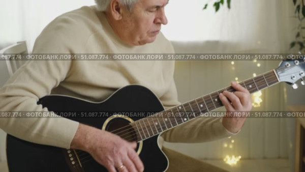 Senior man is playing guitar. Elderly man sitting on the sofa and playing guitar. Portrait of a gray-haired mature man in a sweater and glasses learning to play. Enjoying retirement life at home. Стоковое видео, видеограф Марина Демидюк / Фотобанк Лори