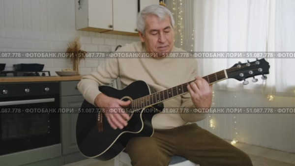 Senior man is playing guitar. Elderly man sitting on the sofa and playing guitar. Portrait of a gray-haired mature man in a sweater and glasses learning to play. Enjoying retirement life at home. Стоковое видео, видеограф Марина Демидюк / Фотобанк Лори