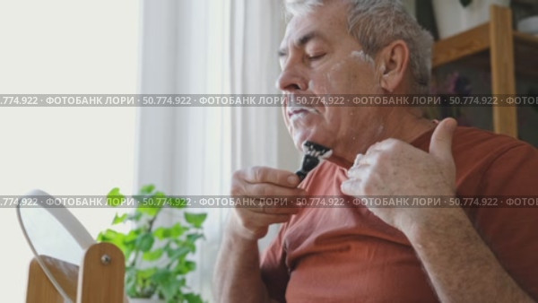 Senior Man shaving his face. Old male shaves beard and mustache with shaver. Elderly gray haired man looking at the mirror, applies shaving cream foam and using razor. Skin hygiene. Daily body care. Стоковое видео, видеограф Марина Демидюк / Фотобанк Лори