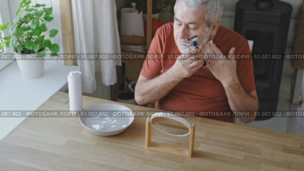 Senior Man shaving his face. Old male shaves beard and mustache with shaver. Elderly gray haired man looking at the mirror, applies shaving cream foam and using razor. Skin hygiene. Daily body care. Стоковое видео, видеограф Марина Демидюк / Фотобанк Лори
