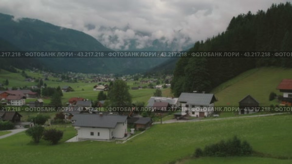 Serene village with houses scattered on a green hillside, surrounded by forested mountains and a cloudy sky, creating a peaceful atmosphere. Стоковое видео, видеограф Igor Tichonow / Фотобанк Лори