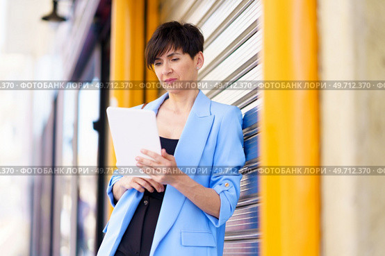 Serious entrepreneur in formal clothes standing by shining metallic gate while browsing tablet and looking at screen outdoors. Serious woman with tablet by metallic gate on street. Стоковое фото, агентство Ingram Publishing / Фотобанк Лори