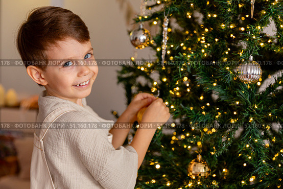 Side view of a cute happy little boy decorating a Christmas tree in the living room at home. Merry Christmas and happy New Year winter holidays. High-quality photo. Стоковое фото, фотограф Ирина Михайличенко / Фотобанк Лори