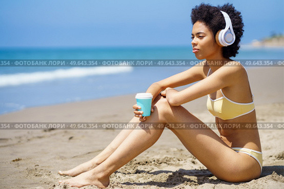Side view of dreamy young African American female tourist with Afro hair, in bikini drinking takeaway coffee and looking away while listening to music in headphones on sandy seashore. Black woman in headphones drinking takeaway beverage while relaxing on beach. Стоковое фото, агентство Ingram Publishing / Фотобанк Лори