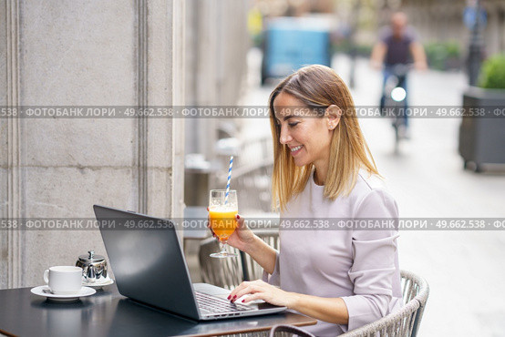 Side view of positive adult self employed lady with blond hair in casual clothes, drinking glass of fresh juice and smiling while working remotely on laptop in street cafe. Happy female freelancer working on netbook in outdoor cafeteria. Стоковое фото, агентство Ingram Publishing / Фотобанк Лори