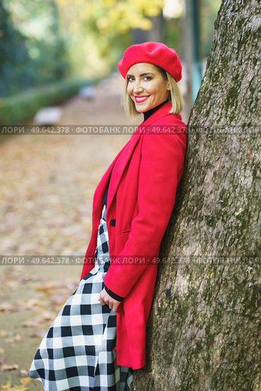 Side view of smiling female wearing red outerwear and checkered shirt leaning on tree in autumn park and smiling at camera. Charming woman standing near tree trunk in park. Стоковое фото, агентство Ingram Publishing / Фотобанк Лори