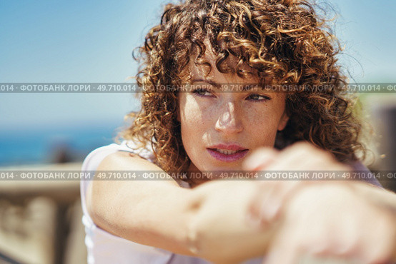 Side view of young female with curly hair looking away thoughtfully while leaning on fence against blurred background in daytime. Young woman with curly hair and freckles on face. Стоковое фото, агентство Ingram Publishing / Фотобанк Лори