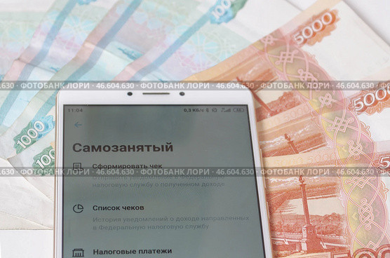 Смартфон с открытым банковским приложением -  личным кабинетом самозанятого (плательщиков налога на профессиональный доход) на фоне пятитысячных и тысячных  купюр. Стоковое фото, фотограф Snezka / Фотобанк Лори