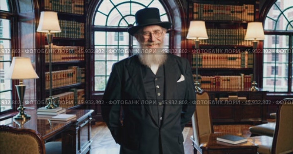 Smiling elderly intelligent man with beard in black suit and hat in classic antique living room with many books. Стоковое AI-видео, аниматор Александр Довянский / Фотобанк Лори
