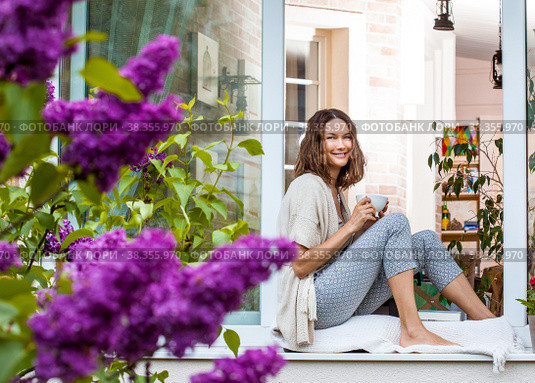 Smiling middle-aged woman drinking tea or coffee on the windowsill. Стоковое фото, фотограф Astroid / Фотобанк Лори