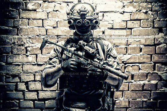 Special forces fighter standing against a brick wall, armed with thermal imaging device on helmet and submachine gun. Стоковое фото, агентство ТАСС / Фотобанк Лори